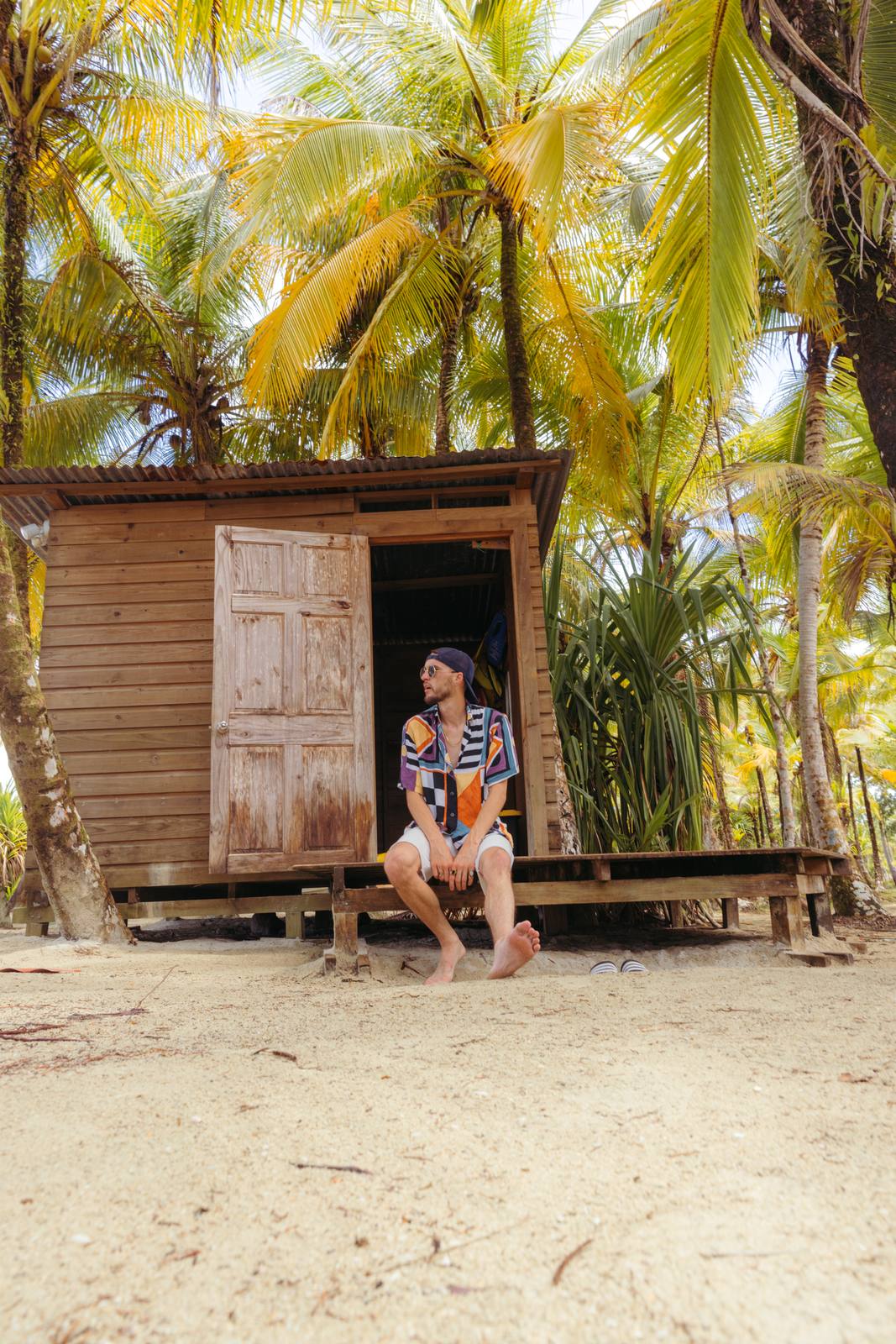 Guest relaxing outside a rustic wooden beach cabin shaded by coconut palms